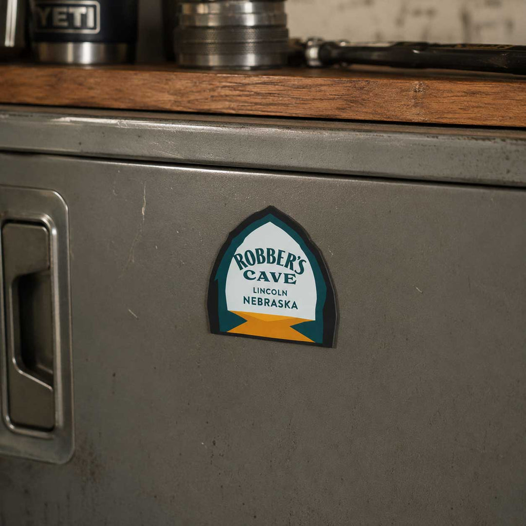 A small arched magnet reading 'Robber's Cave Lincoln Nebraska' applied to the front of a gray metal cabinet, with a wooden surface and metal containers visible above.