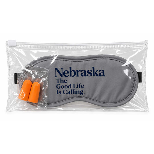 A gray sleep mask with navy blue text reading 'Nebraska The Good Life Is Calling.' inside a clear zippered plastic sleeve, with two orange earplugs visible, on a white background.