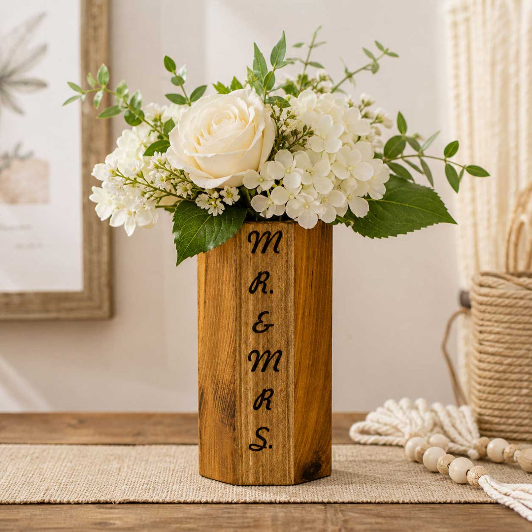 Wooden vase with engraved initials 'M R & MRS' holding a floral arrangement on a table.