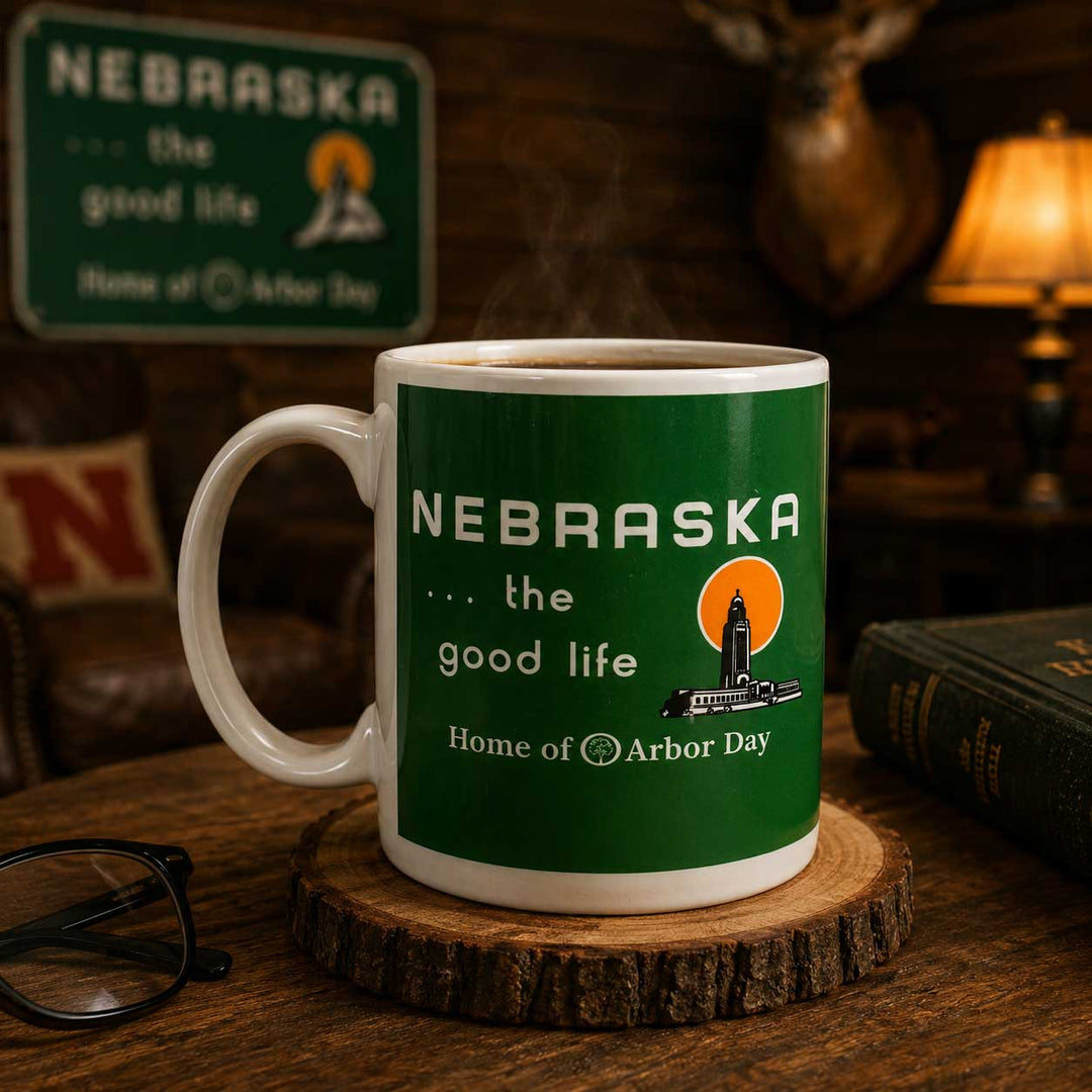 A white ceramic mug with a green Nebraska-themed graphic sits on a rustic wood slice coaster on a wooden table. The mug features the text 'NEBRASKA .