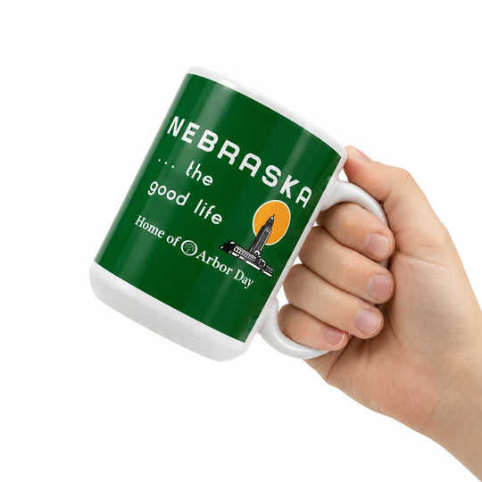 A hand holding a white ceramic mug with a dark green wraparound print that reads 'NEBRASKA .