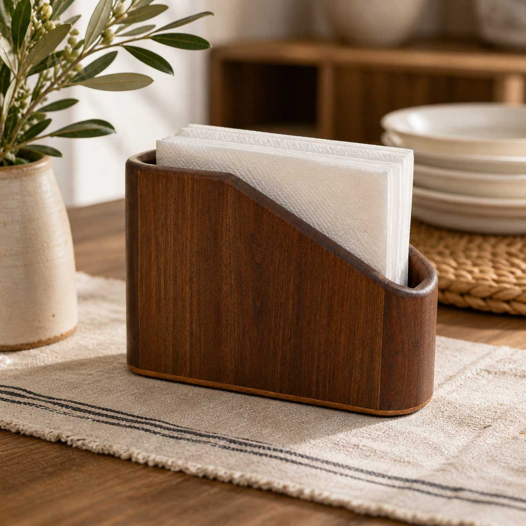Wooden napkin holder with white napkins on a table with a plant and bowls in the background.