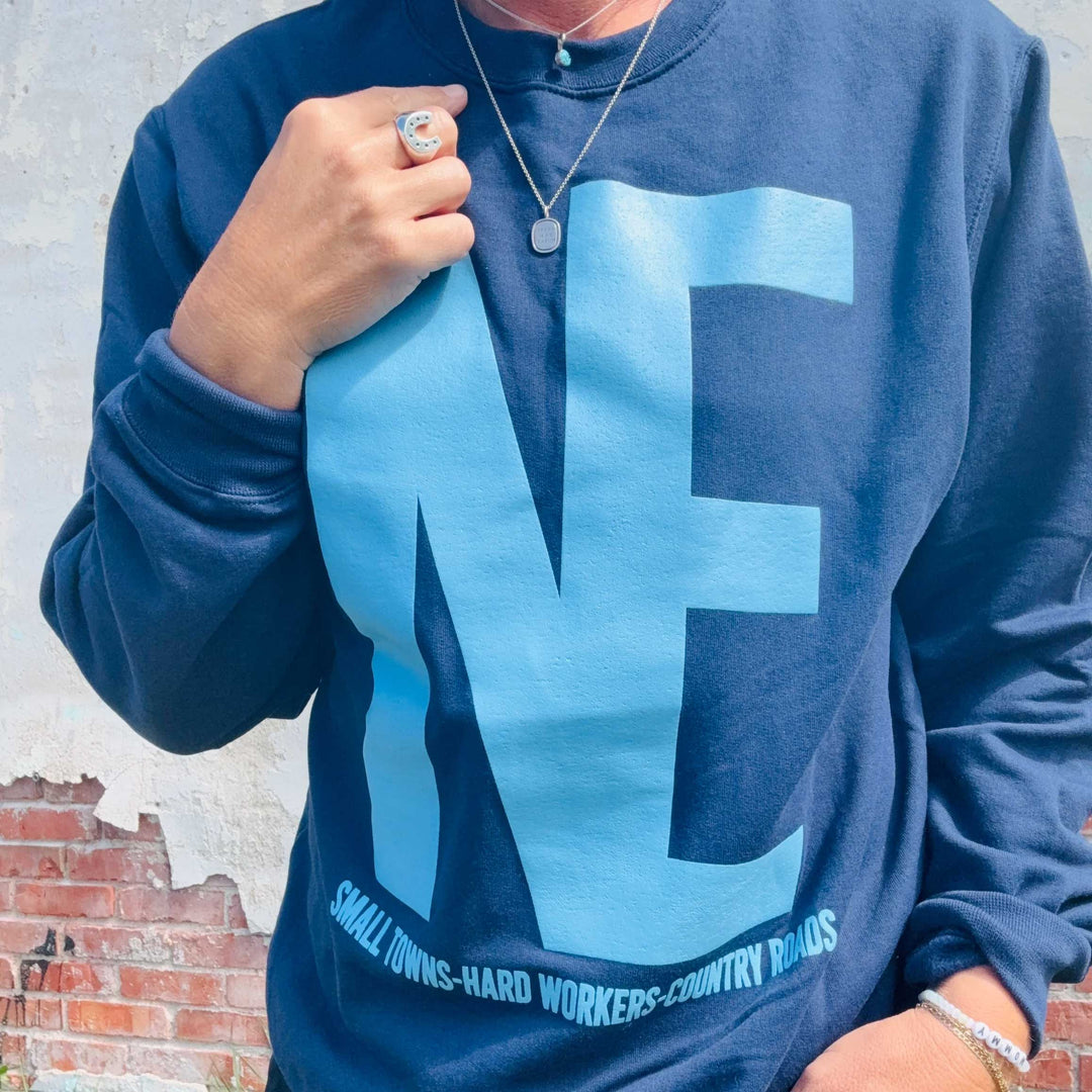 A person wearing a navy blue sweatshirt with a large light blue 'NE' graphic and the text 'SMALL TOWNS-HARD WORKERS-COUNTRY ROADS' printed along the bottom. The person is also wearing a silver necklace and ring, standing against a weathered brick and concrete wall.