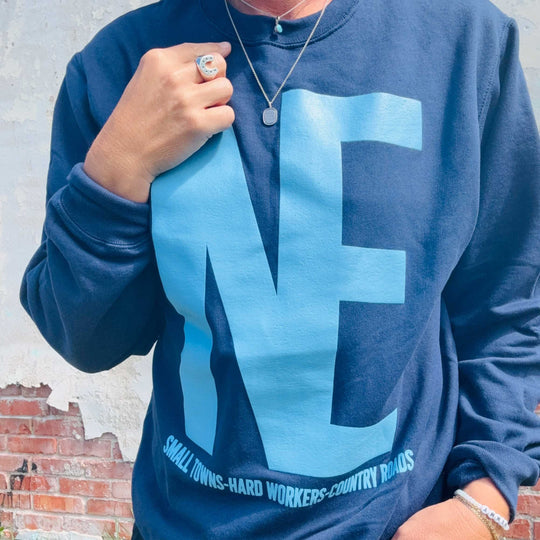 A person wearing a navy blue sweatshirt with a large light blue 'NE' graphic and the text 'SMALL TOWNS-HARD WORKERS-COUNTRY ROADS' printed along the bottom. The person is also wearing a silver necklace and ring, standing against a weathered brick and concrete wall.
