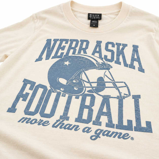 A close-up of a cream-colored t-shirt featuring a distressed blue graphic that reads 'NEBRASKA FOOTBALL more than a game' with a football helmet illustration. The neck label shows the brand 'LUX DESIGN'.