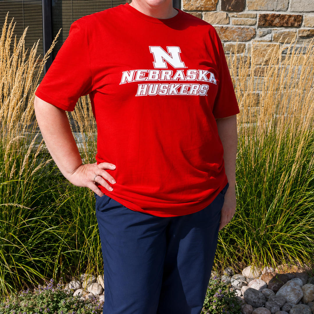 Person wearing a red 'Nebraska Huskers' t-shirt in front of a stone wall and plants.