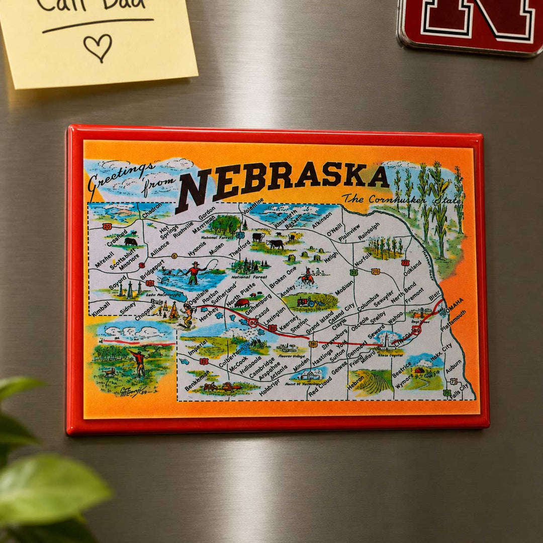A rectangular refrigerator magnet featuring a colorful illustrated map of Nebraska, shown attached to a stainless steel refrigerator. The magnet has a red border, an orange background, and a white map area with various landscape and cultural details.