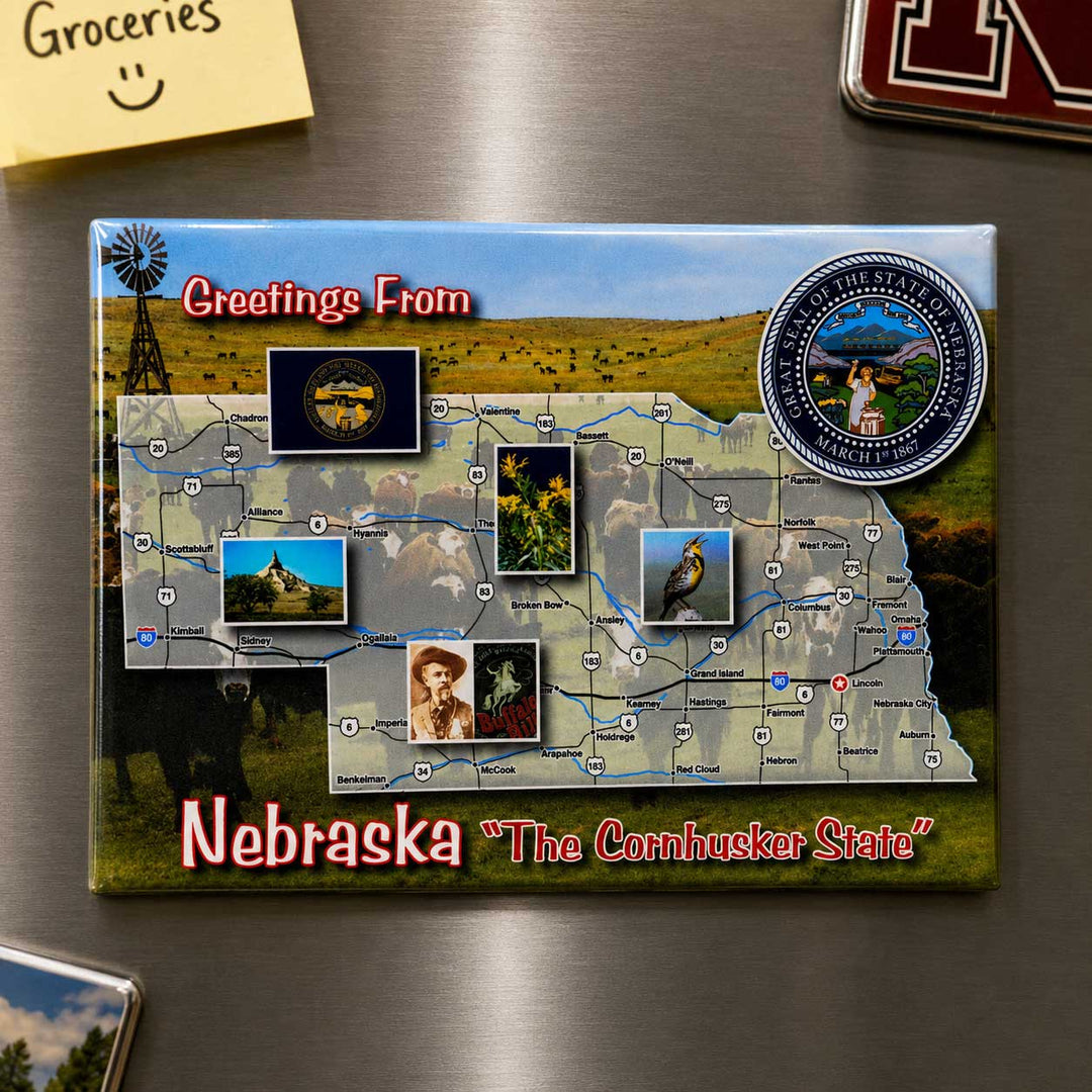 A rectangular Nebraska-themed magnet or plaque on a brushed metal surface, featuring a map of Nebraska with the text 'Greetings From' and 'Nebraska "The Cornhusker State"'. The design includes a blue sky, green fields, a windmill, circular state seal, and small inset photos.