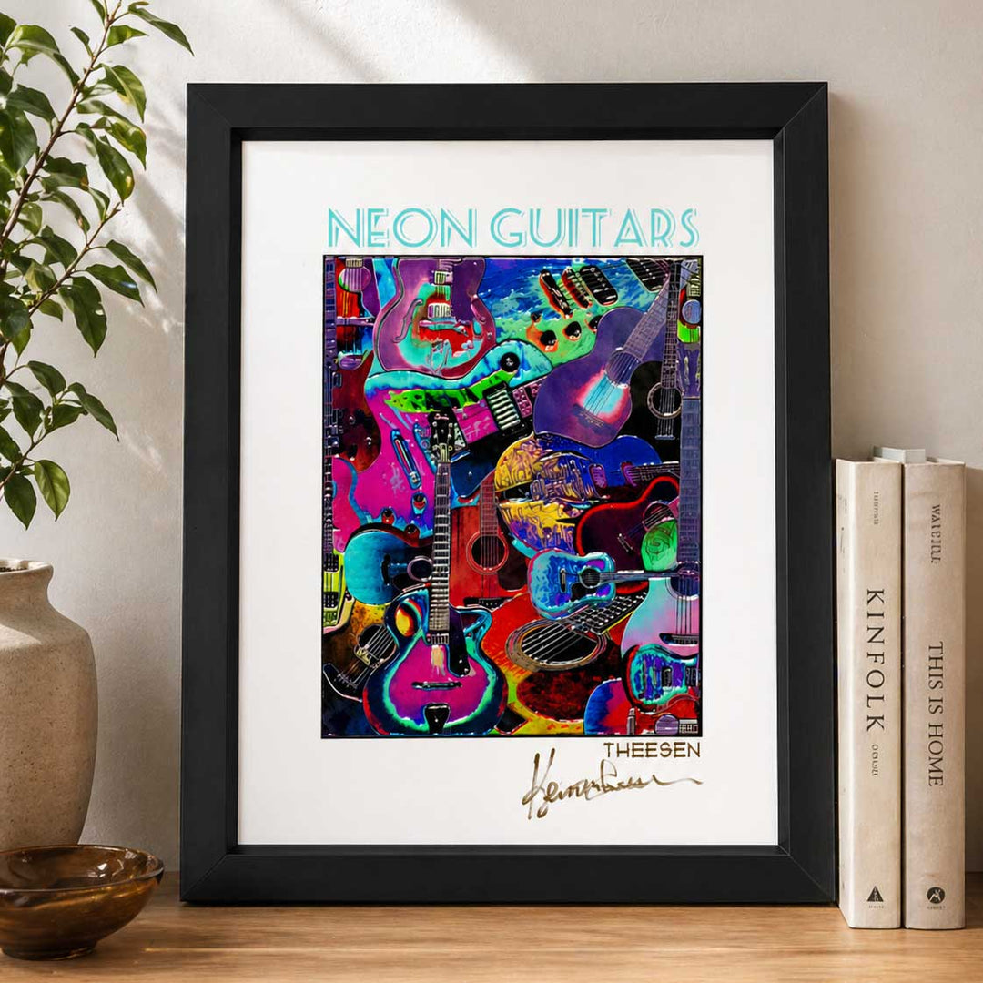 Framed artwork titled 'Neon Guitars' by Theesen on a wooden surface with books and a plant.