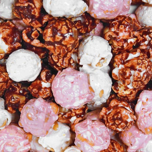 A close-up pile of gourmet popcorn mixed with white chocolate and pink chocolate pieces, showing glossy caramel-coated kernels alongside smooth white and pink coated popcorn balls.
