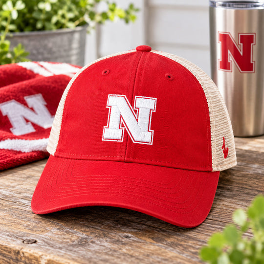 A red and beige trucker hat with a white embroidered Nebraska 'N' logo on the front, displayed on a rustic wooden table with a matching red towel and a stainless steel tumbler in the background.