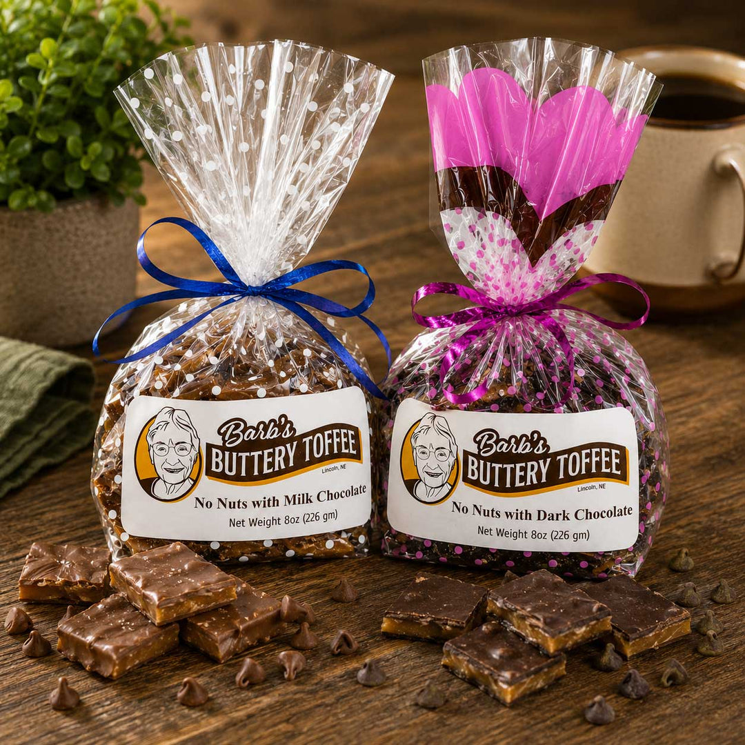 Two bags of Barb's Buttery Toffee presented on a wooden table, with loose toffee pieces scattered in front. One bag contains toffee with nuts and is tied with a blue ribbon, while the other contains toffee without nuts and is tied with a purple ribbon.