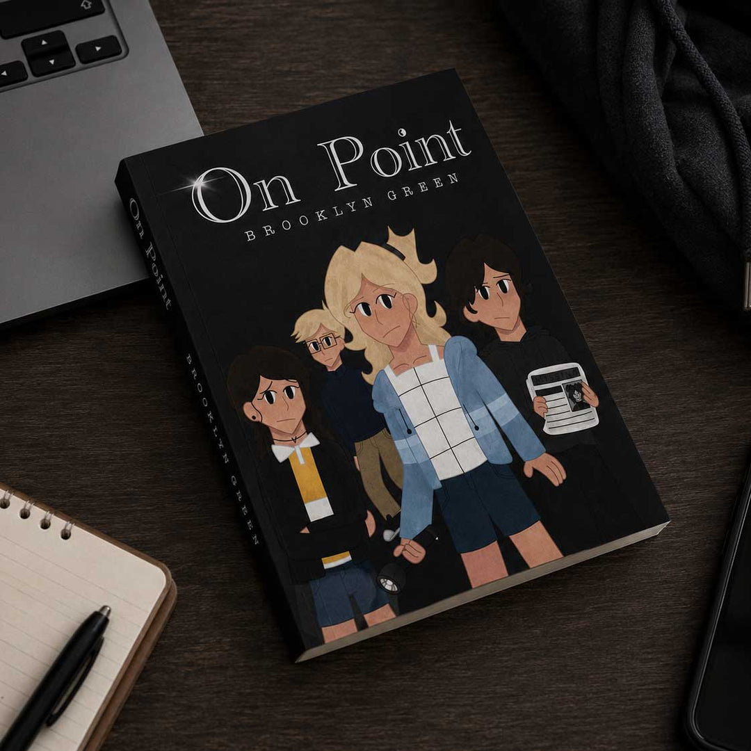 A hardcover book titled "On Point: Brooklyn Green" lies on a dark wooden desk, surrounded by a laptop, notebook, pen, and dark fabric. The cover is black with illustrated characters and white title text.