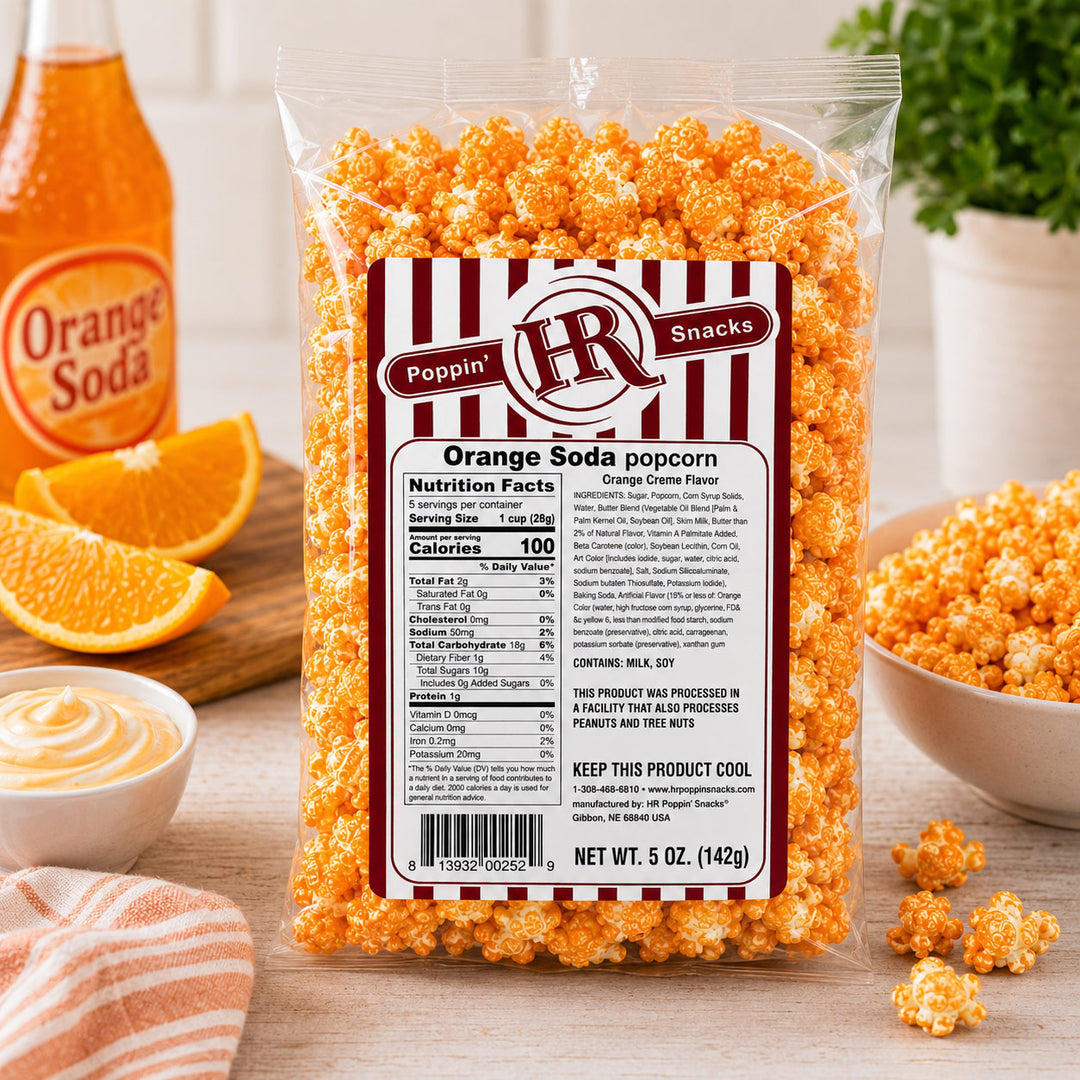 A clear plastic bag of Poppin' Snacks Orange Soda popcorn sits on a wooden table, surrounded by orange slices, a bowl of popcorn, a small dish of dip, and an orange soda bottle in the background.