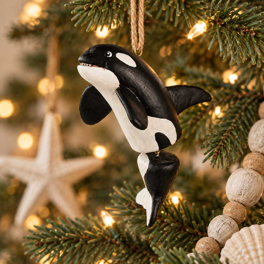 A hand-painted wooden orca fish ornament hanging from a jute string on a decorated Christmas tree with warm lights, a wooden star, and a bead garland.