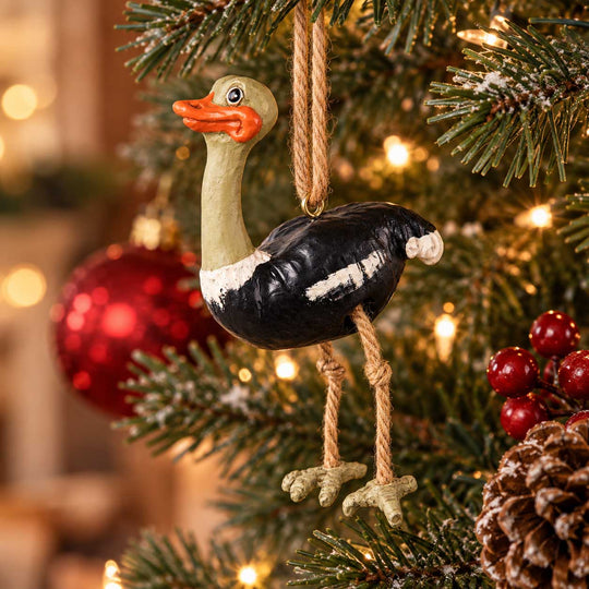 A painted ostrich Christmas ornament hanging from a tree branch with a jute loop, surrounded by a decorated Christmas tree with lights, red ornaments, pinecones, and berries.