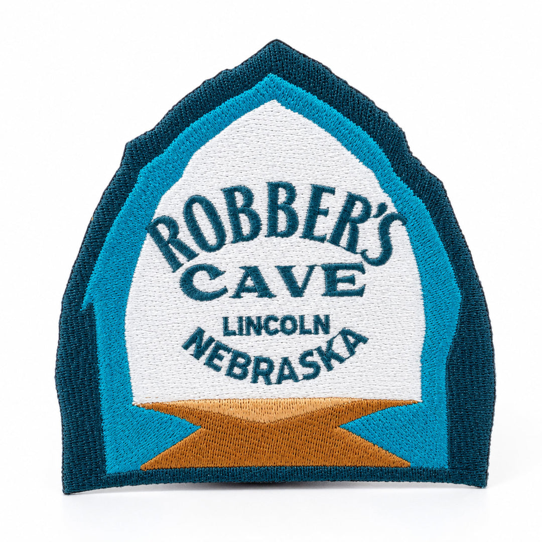 An embroidered patch shaped like a tombstone, featuring the text 'ROBBER'S CAVE LINCOLN NEBRASKA' in dark blue embroidery on a white background, surrounded by a teal border and a gold star at the bottom, set against a plain white background.