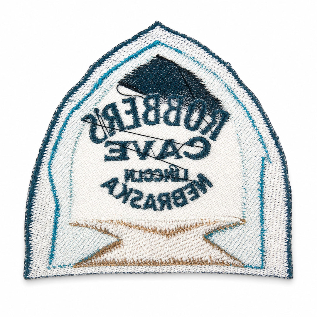 An embroidered arch-shaped patch featuring the words 'Robbers Cave Lincoln Nebraska' in dark blue text over a white background, with a dark blue house silhouette at the top and a tan/gold banner at the bottom. A black thread ball is threaded through the center.
