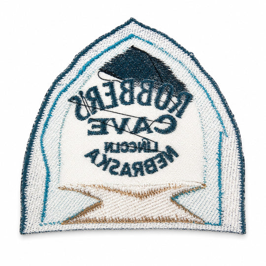 An embroidered arch-shaped patch featuring the words 'Robbers Cave Lincoln Nebraska' in dark blue text over a white background, with a dark blue house silhouette at the top and a tan/gold banner at the bottom. A black thread ball is threaded through the center.