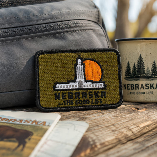 A rectangular embroidered patch featuring the Nebraska state capitol and an orange sun on an olive green background, placed on a wooden surface with a gray bag, a mug, and a Nebraska-themed card nearby.