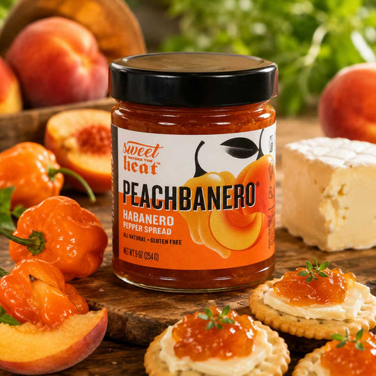 A jar of Sweet Heat Peachbanero Habanero Pepper Spread is shown on a wooden board with sliced peaches, habanero peppers, crackers topped with spread, and a block of cheese in a styled food setting.