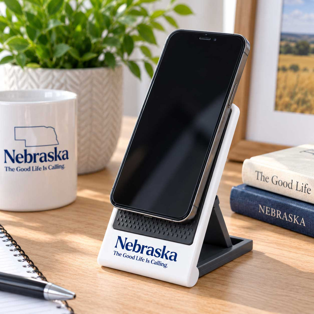 A white and black Nebraska-themed phone stand holding a smartphone, set on a wooden desk with a mug, plant, books, and framed picture in the background.