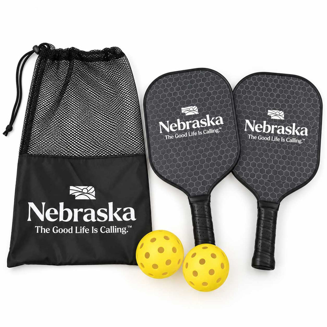 A set of pickleball paddles with two yellow pickleballs and a black drawstring bag, all featuring the Nebraska logo and 'The Good Life Is Calling' slogan on a white background.