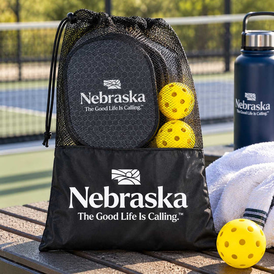 A black mesh drawstring bag with a large front pocket printed with the Nebraska logo and slogan, shown on a tennis court. The bag contains a black paddle, two yellow perforated balls, and a white towel.