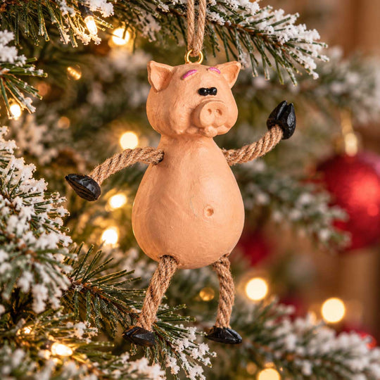 A pig-shaped Christmas ornament hanging from a decorated Christmas tree. The ornament has a light peach-pink body, rope arms and legs, and black hooves and nose details, set against a festive background of green tree branches, white snow-like dusting, and warm yellow lights.