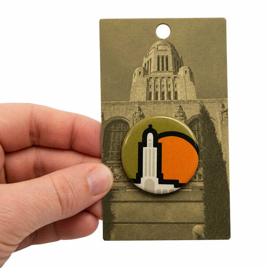 A hand holds a rectangular display card featuring a round pin or badge design. The badge has a stylized white building silhouette over an olive green and orange background, with a sepia-toned building photo printed on the card behind it.