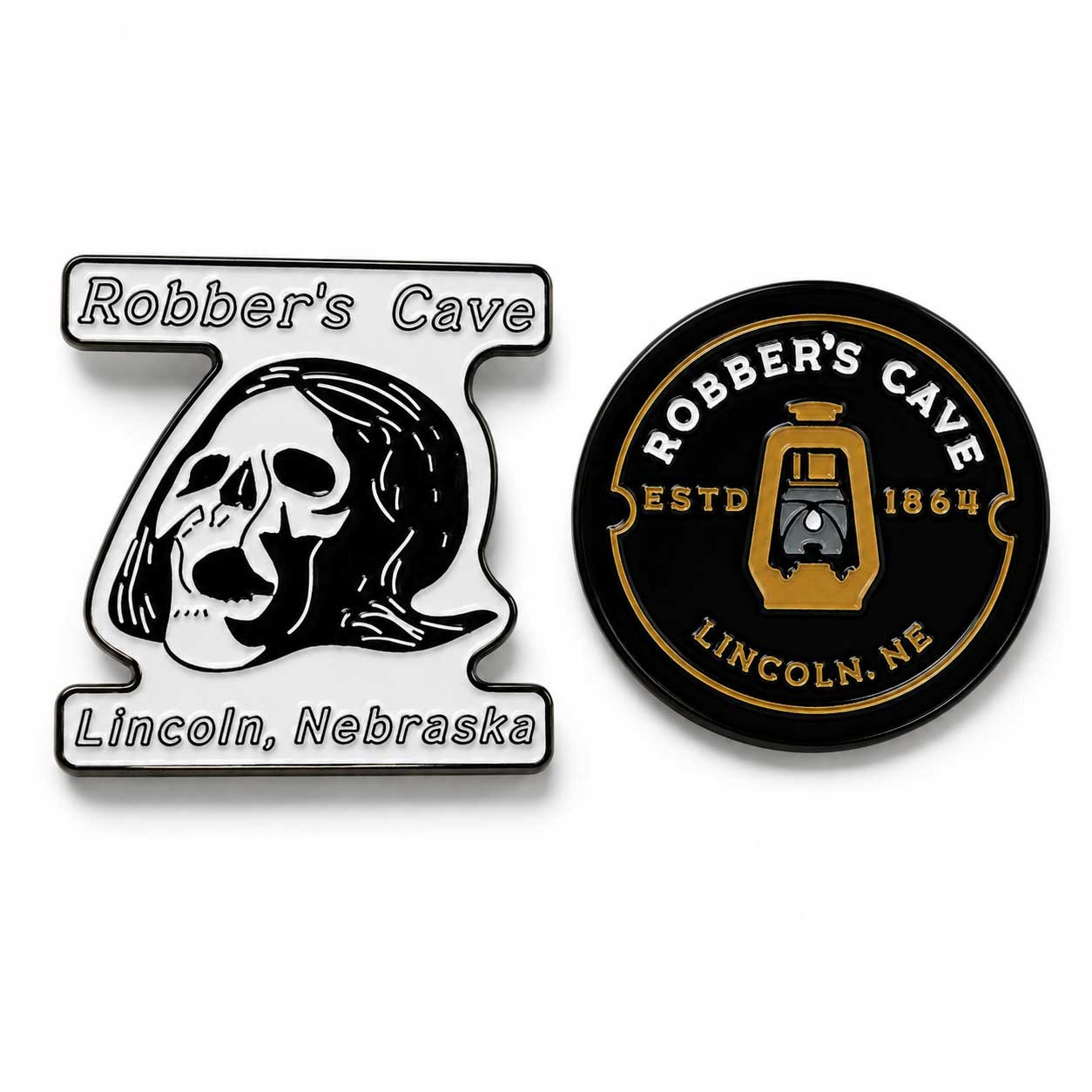 Two enamel pins displayed side-by-side on a white background. The left pin is shaped like the number 7, featuring a black and white skull illustration with the text 'Robber's Cave' and 'Lincoln, Nebraska'.