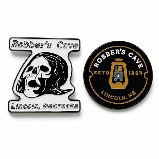 Two enamel pins displayed side-by-side on a white background. The left pin is shaped like the number 7, featuring a black and white skull illustration with the text 'Robber's Cave' and 'Lincoln, Nebraska'.