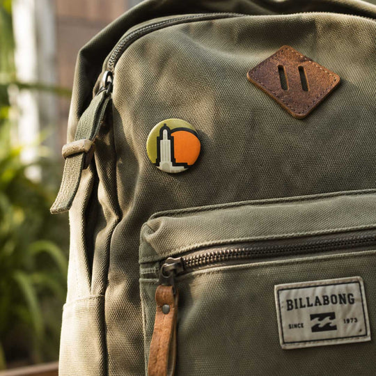 Close-up of an olive green Billabong canvas backpack with a brown leather patch, a circular multicolor pin, and a Billabong brand label on the front pocket.