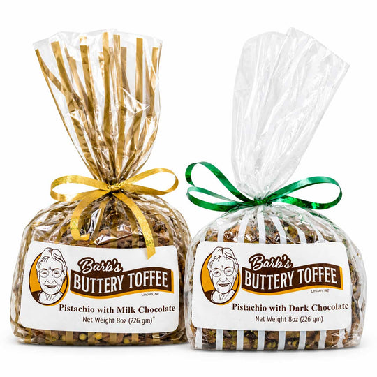 Two bags of Barb's Buttery Toffee on a white background. The left bag is labeled 'Pistachio with Milk Chocolate' and tied with a gold ribbon, while the right bag is labeled 'Pistachio with Dark Chocolate' and tied with a green ribbon.