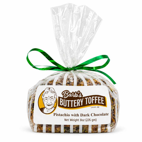 A 8 oz bag of Barb's Buttery Toffee Pistachio with Dark Chocolate, wrapped in clear cellophane with a green ribbon bow and a white label featuring the brand logo.