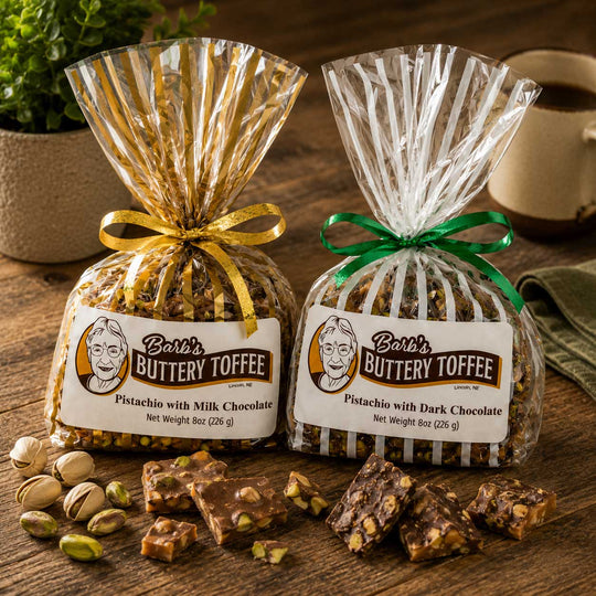 Two bags of Barb's Buttery Toffee, one with gold foil wrapping and one with clear wrapping and a green ribbon, displayed on a wooden table with pieces of toffee and pistachios scattered in front.