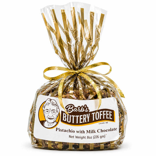 A 8 oz bag of Barb's Buttery Toffee Pistachio with Milk Chocolate, wrapped in clear cellophane with gold foil streamers and a gold ribbon bow, on a white background.