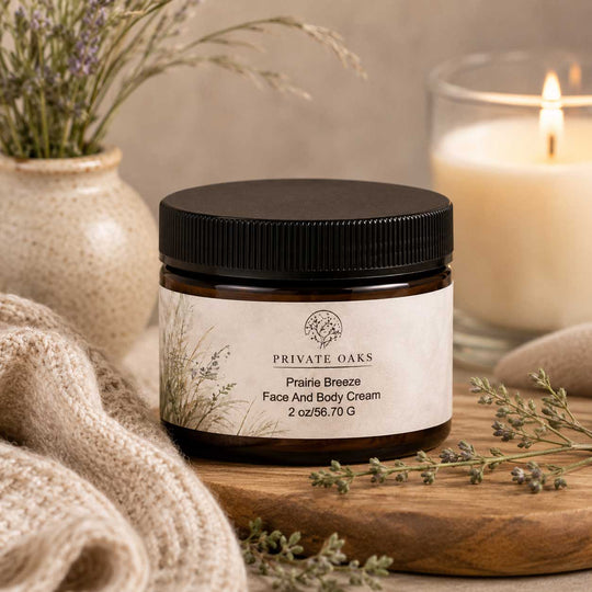 A jar of Private Oaks Prairie Breeze Face And Body Cream sits on a wooden board surrounded by dried flowers, a knitted beige cloth, and a softly lit candle in the background.