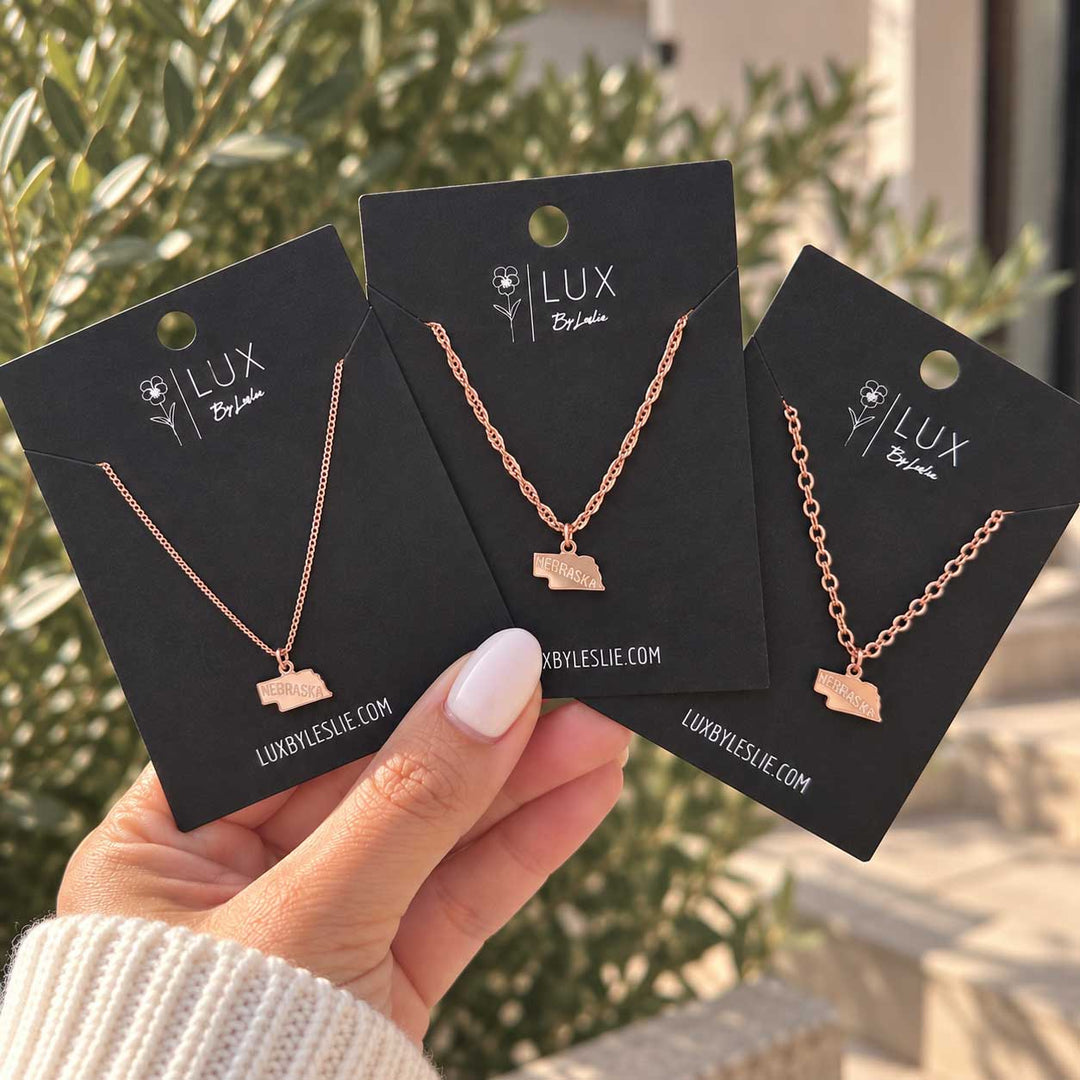 Three rose gold necklaces with small state-of-California pendants are displayed on black Lux by Leslie brand cards, held by a hand against a blurred outdoor background of green foliage.