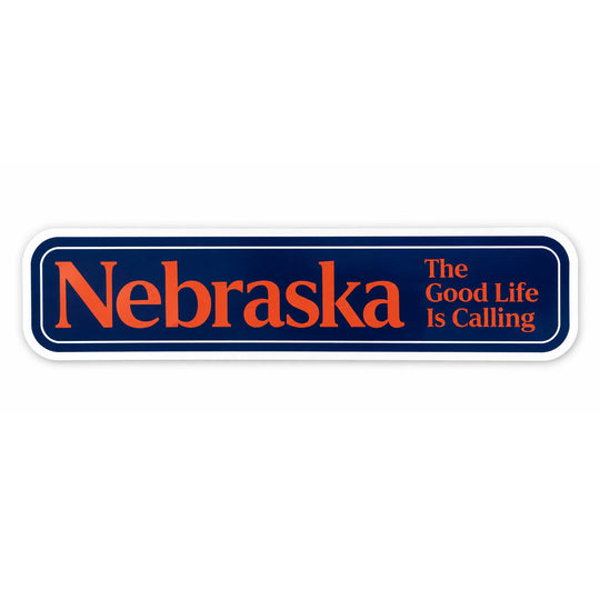 A rectangular bumper sticker with rounded corners featuring the word 'Nebraska' in large orange letters on a dark navy blue background, with the phrase 'The Good Life Is Calling' in smaller orange text to the right. The sticker has a white border and is shown on a plain white background.
