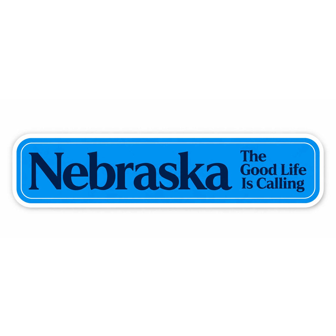 A rectangular blue sticker or sign with rounded corners, featuring the word 'Nebraska' in large dark blue letters and the phrase 'The Good Life Is Calling' in smaller dark blue text on the right. The sticker has a white border and is shown on a plain white background.