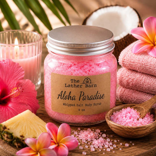 A jar of pink whipped salt body scrub by The Lather Barn, labeled 'Aloha Paradise,' styled on a wooden surface with pink towels, hibiscus flowers, pineapple, a candle, and a small bowl of bath salts.