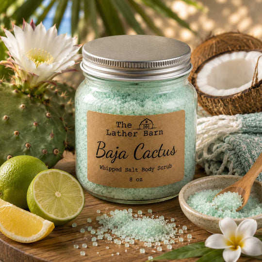 A jar of The Lather Barn Baja Cactus whipped salt body scrub sits on a wooden surface surrounded by cactus flowers, limes, a halved coconut, a small bowl with a wooden spoon, and scattered salt crystals.
