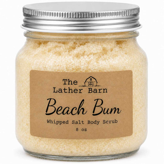 A glass jar of whipped salt body scrub by The Lather Barn, labeled 'Beach Bum'. The scrub inside is a pale, creamy yellow, topped with a silver metal screw-on lid and a brown kraft paper label.