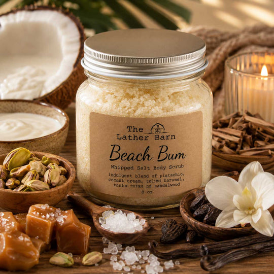 A jar of 'Beach Bum' whipped salt body scrub by The Lather Barn is styled on a rustic wooden table with ingredients like sliced coconut, pistachios, caramel, vanilla, cinnamon, and a lit candle.