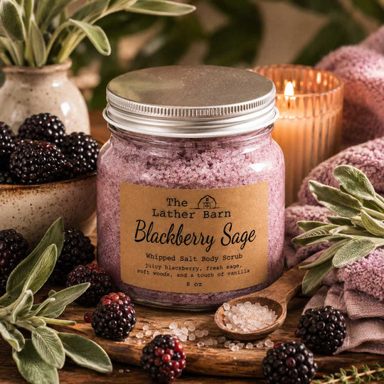 A jar of whipped salt body scrub from The Lather Barn, labeled Blackberry Sage, styled on a rustic wooden surface with blackberries, sage leaves, a lit candle, and bath salts.