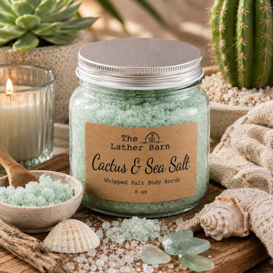 A jar of whipped salt body scrub labeled 'The Lather Barn Cactus & Sea Salt' sits on a wooden surface surrounded by seashells, sea glass, a candle, and bath salts. The product is styled in a beach-inspired setting.
