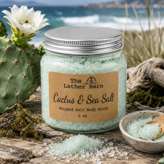 A 5 oz glass jar of 'The Lather Barn' Cactus & Sea Salt Whipped Salt Body Scrub, styled on a rustic wooden surface with a cactus, a small bowl of scrub, and a blurred beach background.