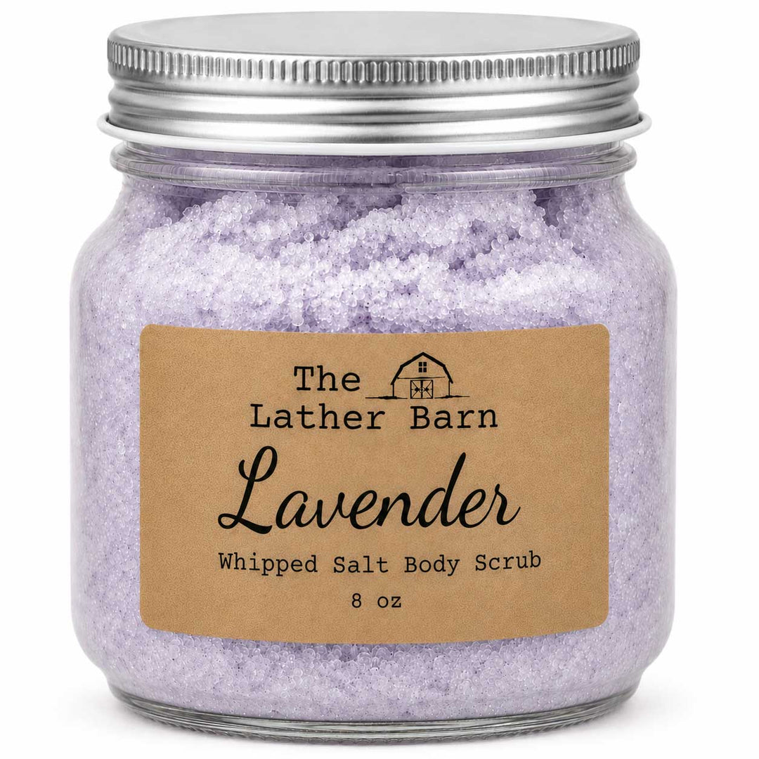 A glass jar filled with lavender whipped salt body scrub, featuring a brown kraft paper label that reads 'The Lather Barn Lavender Whipped Salt Body Scrub 8 oz' and a silver metal screw-top lid, on a white background.