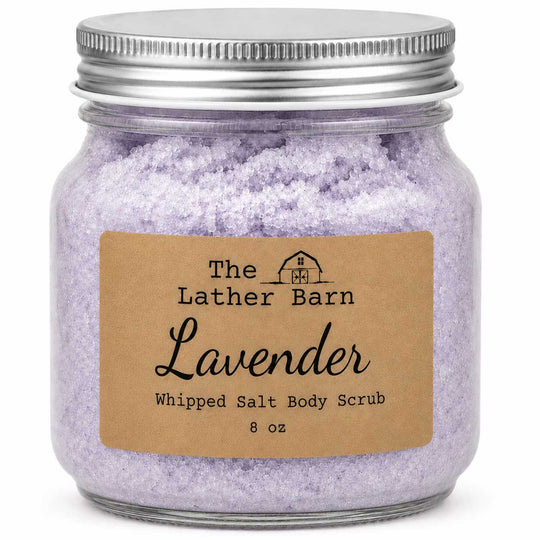 A glass jar filled with lavender whipped salt body scrub, featuring a brown kraft paper label that reads 'The Lather Barn Lavender Whipped Salt Body Scrub 8 oz' and a silver metal screw-top lid, on a white background.